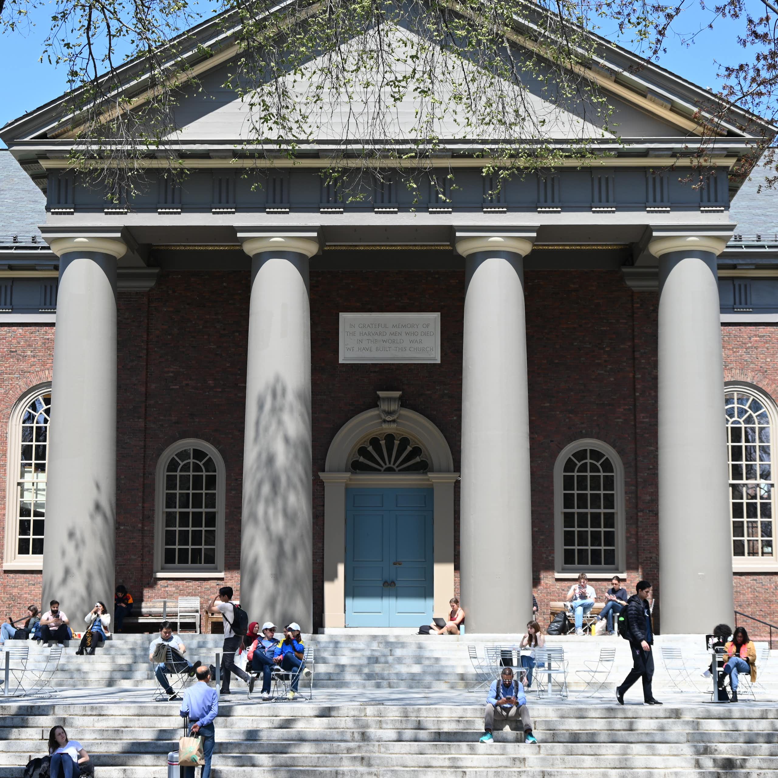 People sit on stairs and on chairs in front of a building on a college campus.