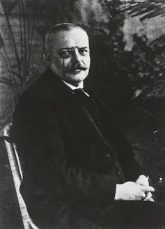 Black and white photo of a seated man with a mustache wearing glasses