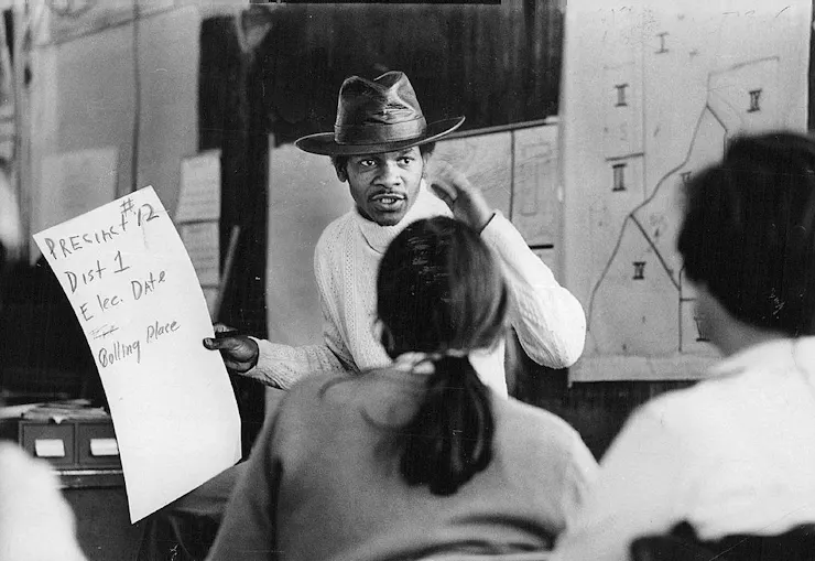 Young Black man wearing a cowboy hat speaks to a group while holding a poster with voting information.