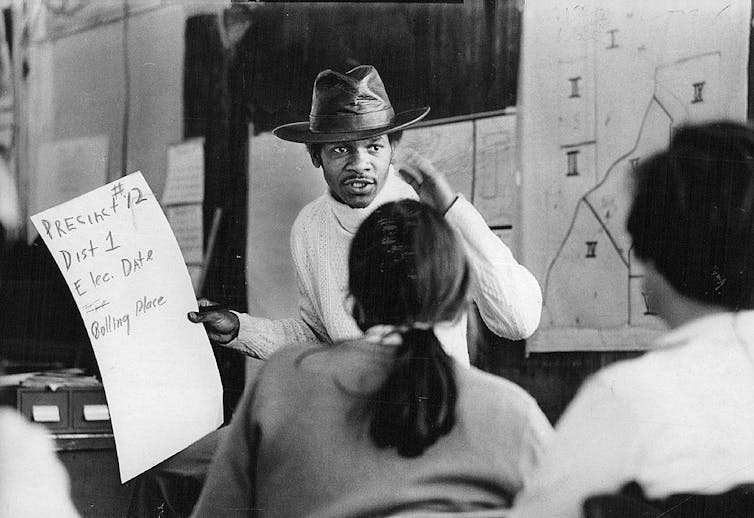 Young Black man wearing a cowboy hat speaks to a group while holding a poster with voting information.