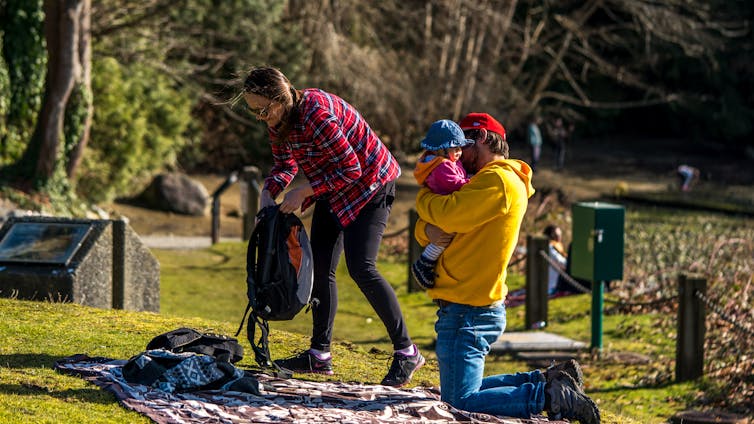 A woman unpacks a backback while a man holds a young girl while kneeling on a blanket in a park