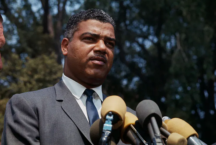 Black man wearing suit stands before microphones.