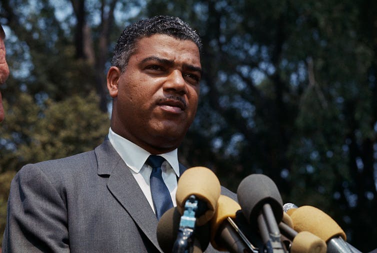 Black man wearing suit stands before microphones.