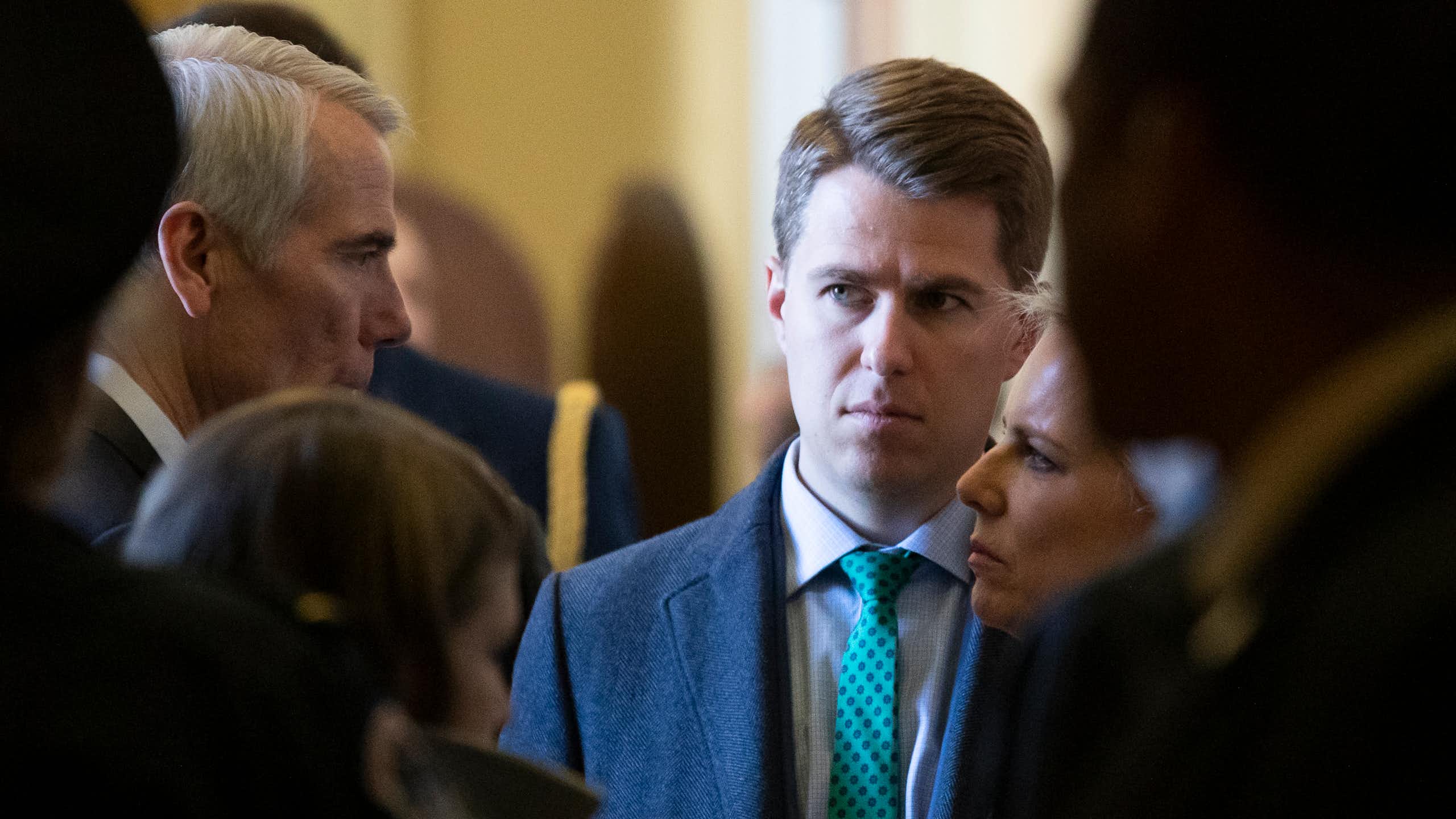 A man in a blue suit and green tie looks at a man speaking.