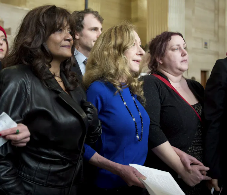 Three women among a crowd speaking to reporters with microphones.