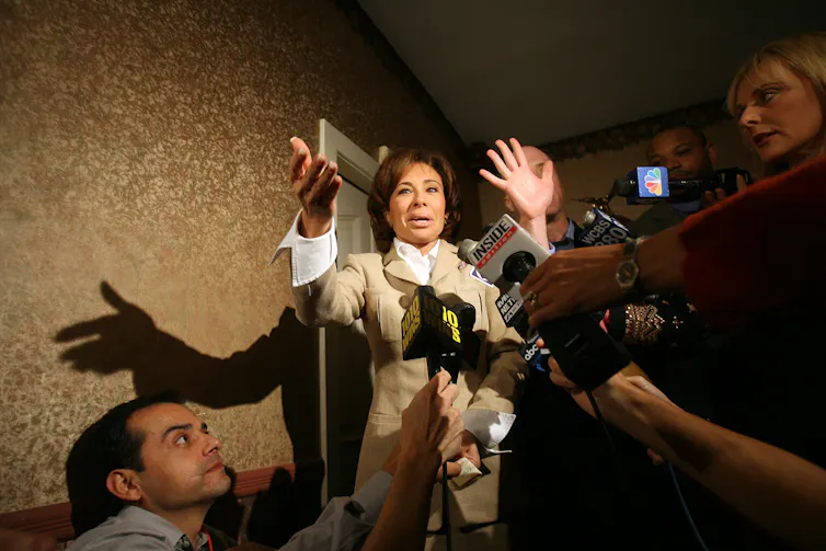 A woman, her hand raised in the air, speaks in from of several news reporters holding microphones.