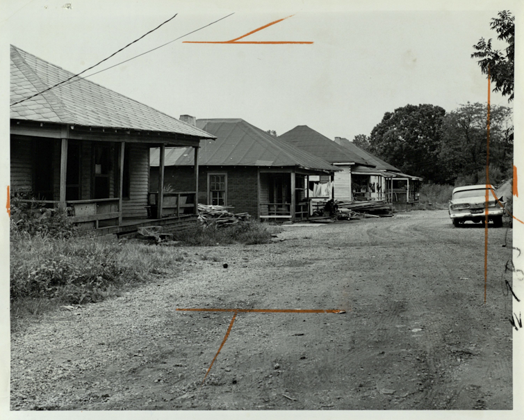 Black and white photo of old, one-story homes along a dirt road.