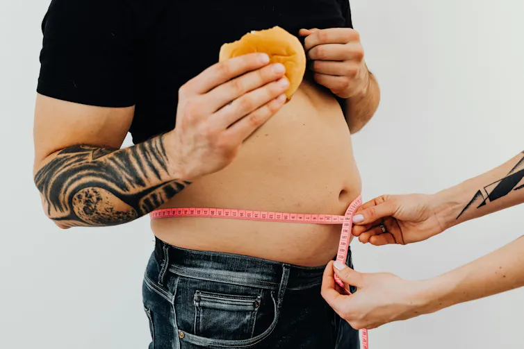 a man with a small belly and tattooed arms eating a burger while hands measure his bare stomach