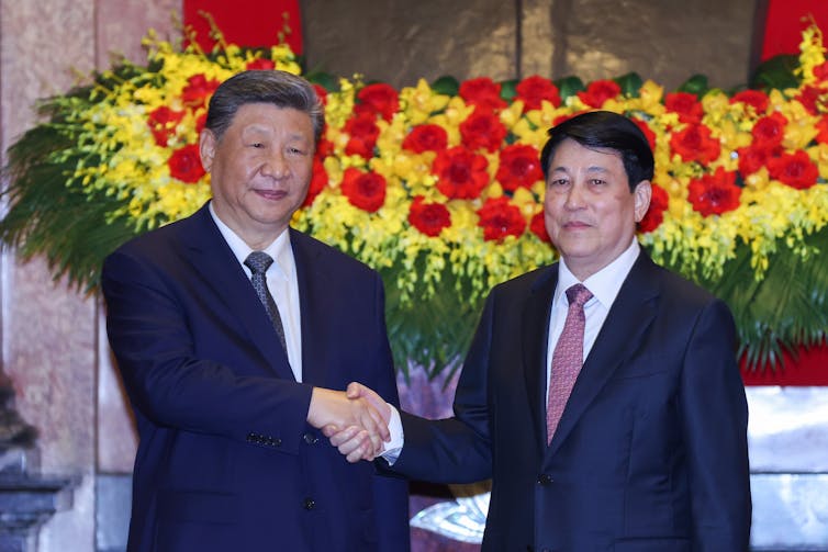 Two Asian men grin while shaking hands in front of a bank of red and yellow flowers.