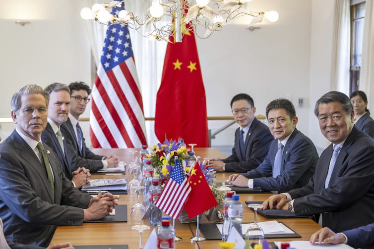 A row of largely stern-faced white men on one side of a table across from a row of smiling Asian men. An American and Chinese flag are in the background.