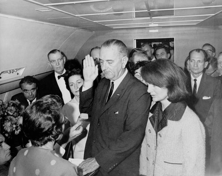 Before a crowd of onlookers, a tall, older man wearing a suit holds his right hand up in the air while his left hand rests on a book.