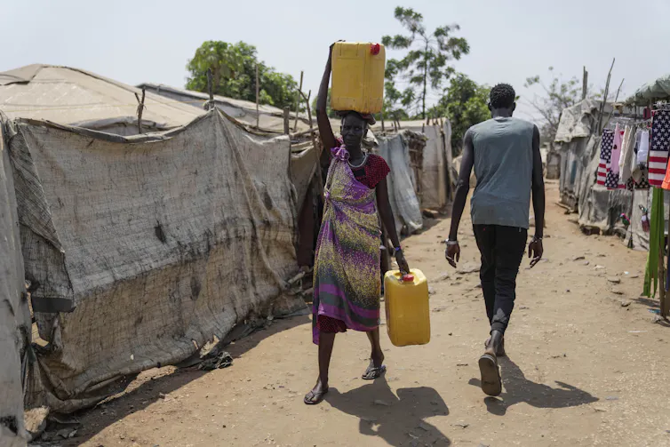 A Black woman carries a container of water on her head and carries another while walking in displacement camp.