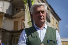 A grey-haired man in a flak jacket stands in front of a bombed building.