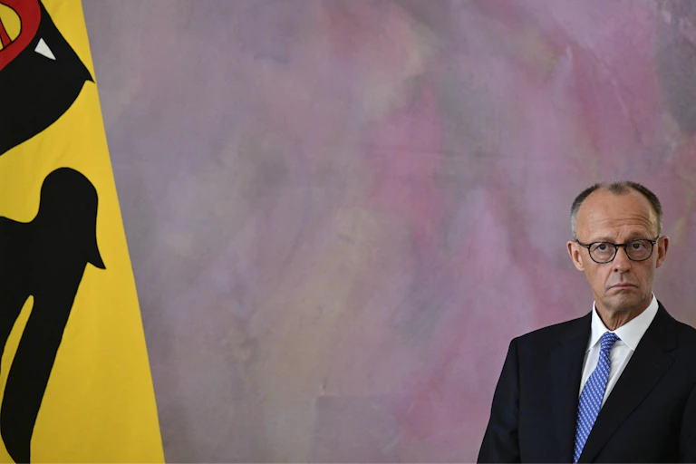 A man in a suit stands next to a yellow flag with a black bird design on it.