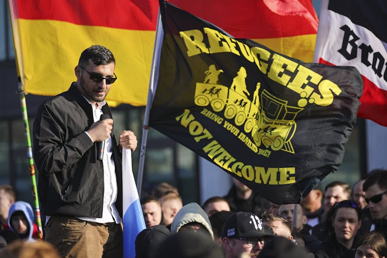 A man in dark glasses stands in front of flags including one stating: 'Refugees not welcome'.