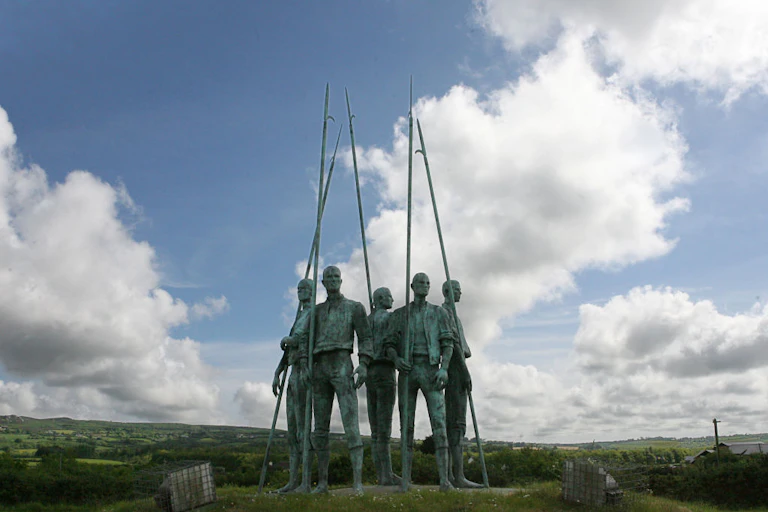 A photo of a green sculpture of several men in a circle, holding spikes, positioned outside under a bright but cloudy sky.