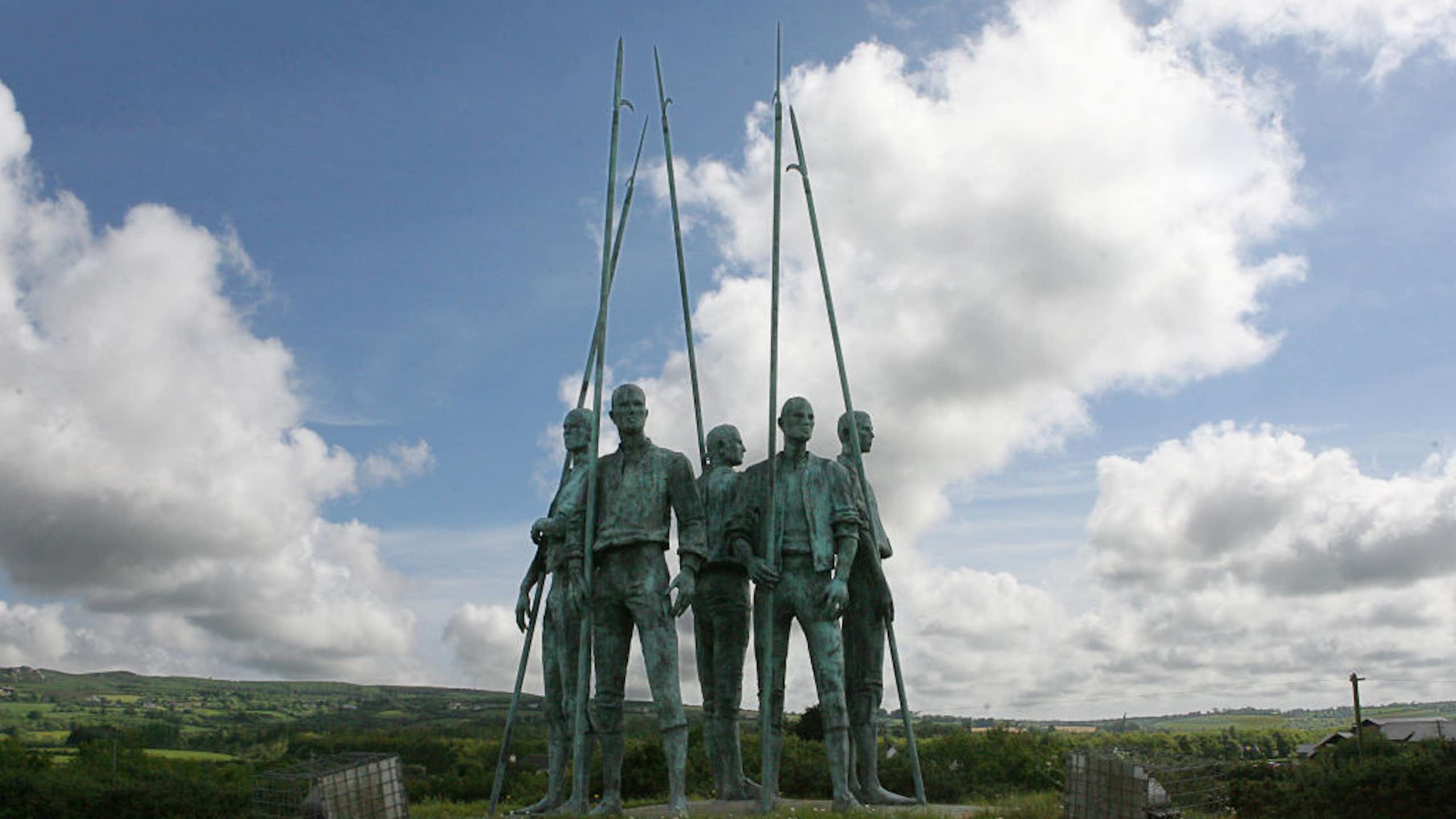 A photo of a green sculpture of several men in a circle, holding spikes, positioned outside under a bright but cloudy sky.
