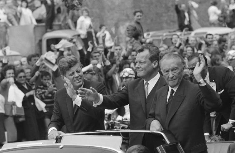 Three men stand on the back of car waving at the crowds.