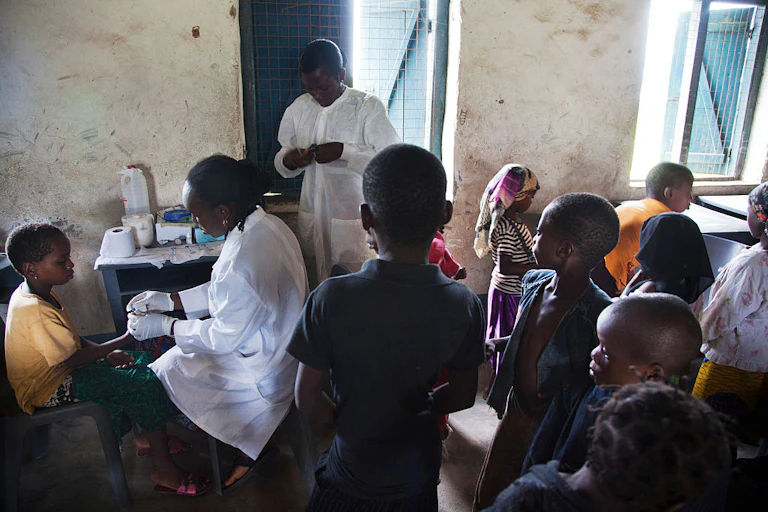 Children milling about a room, waiting to receive a finger prick test from two health care workers
