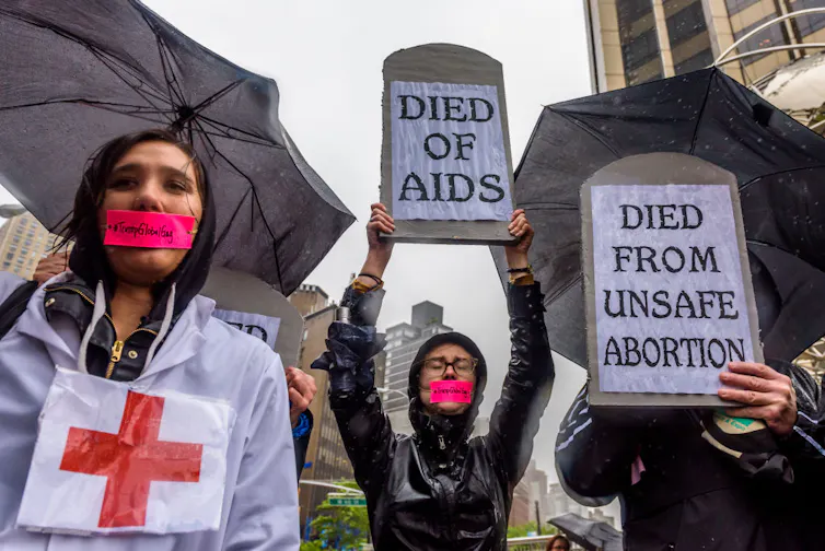 Protectors standing in the rain, holding grave-shaped signs reading 'DIED OF AIDS' and 'DIED FROM UNSAFE ABORTION,' with pink tape reading '#TrumpGlobalGag' over their mouths