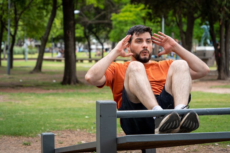 The man sat on the gym equipment in the park.