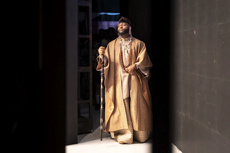 A man stands backstage behind curtains holding a walking stick and regally dressed in a flowing gold robe over loose-fitting gold top and trousers.