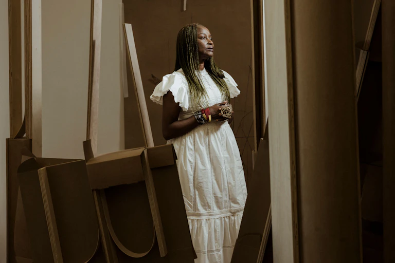 An African woman with long braids stands in a room with art canvasses and easels, her hands held together, pensive in half profile and a long white dress.