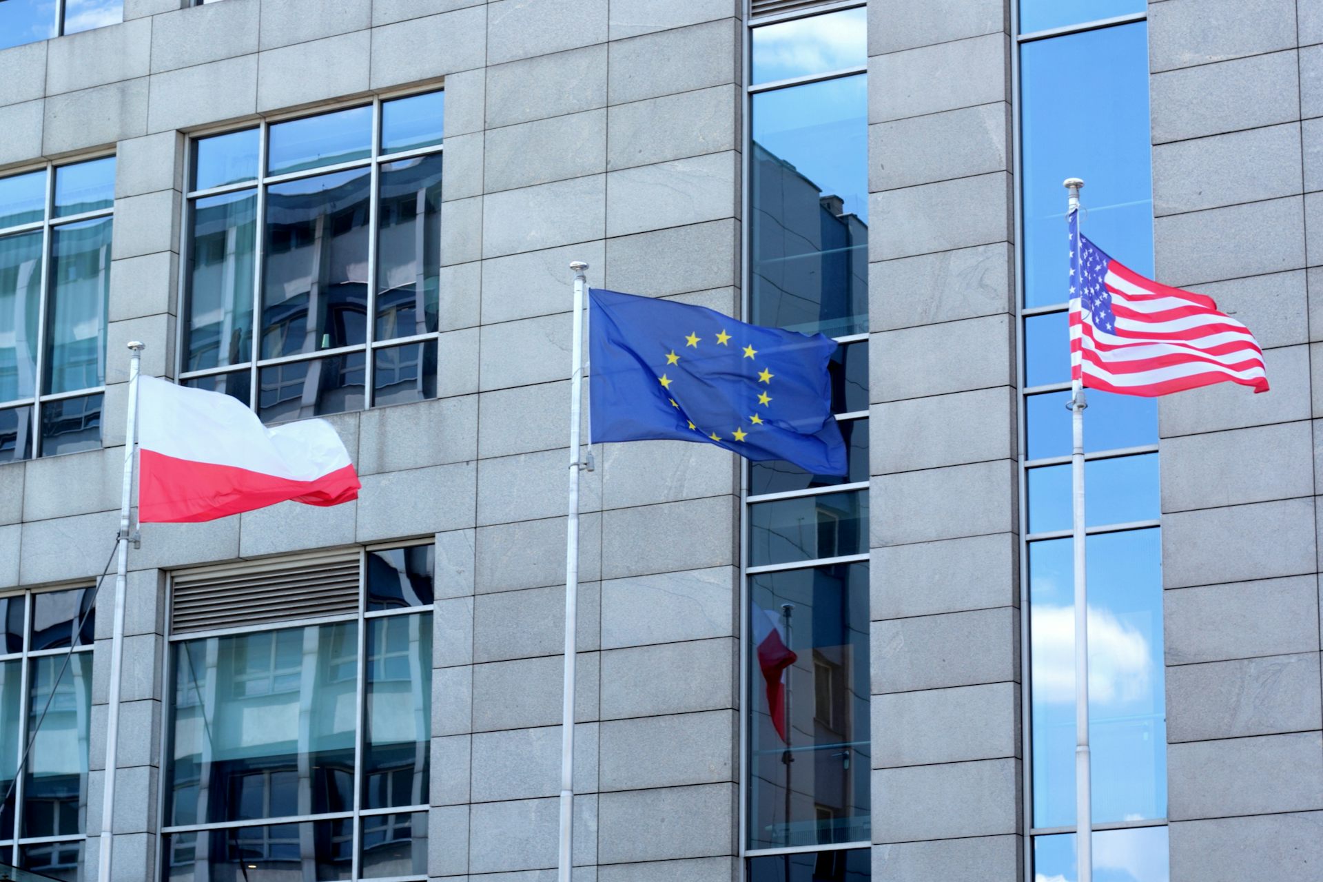 A red and white Polish flag, a blue and yellow EU flag, and a red, white and blue US flag flutter in front of a building in daylight.