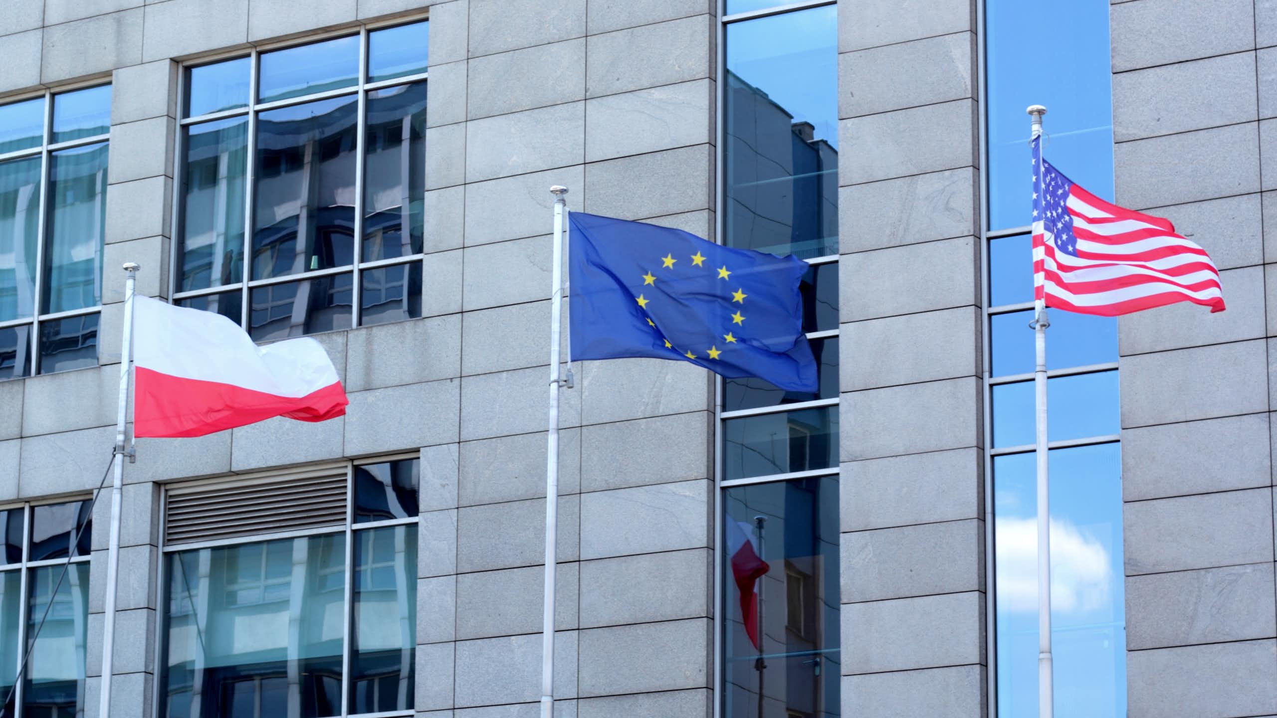 A red and white Polish flag, a blue and yellow EU flag, and a red, white and blue US flag flutter in front of a building in daylight.