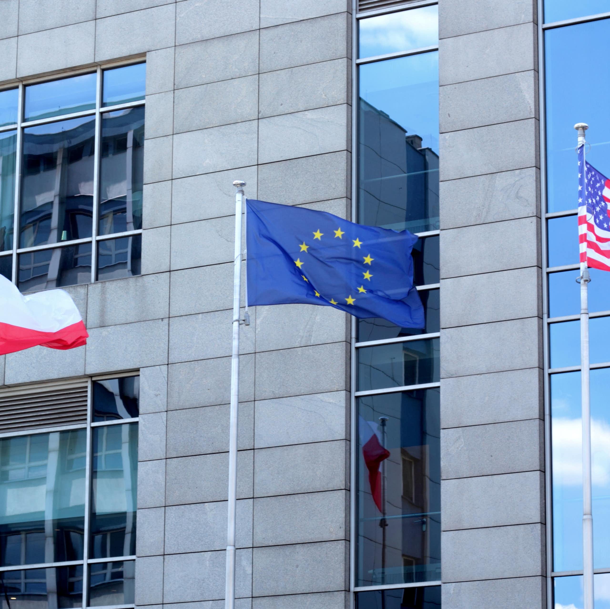 A red and white Polish flag, a blue and yellow EU flag, and a red, white and blue US flag flutter in front of a building in daylight.