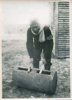 An old black and white photo of an African man bending over and playing drums on a hollowed out log. He's dressed warmly with a scarf and looks directly to camera, eyes grim.
