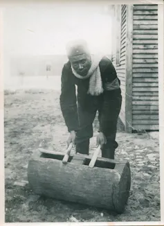 An old black and white photo of an African man bending over and playing drums on a hollowed out log. He's dressed warmly with a scarf and looks directly to camera, eyes grim.