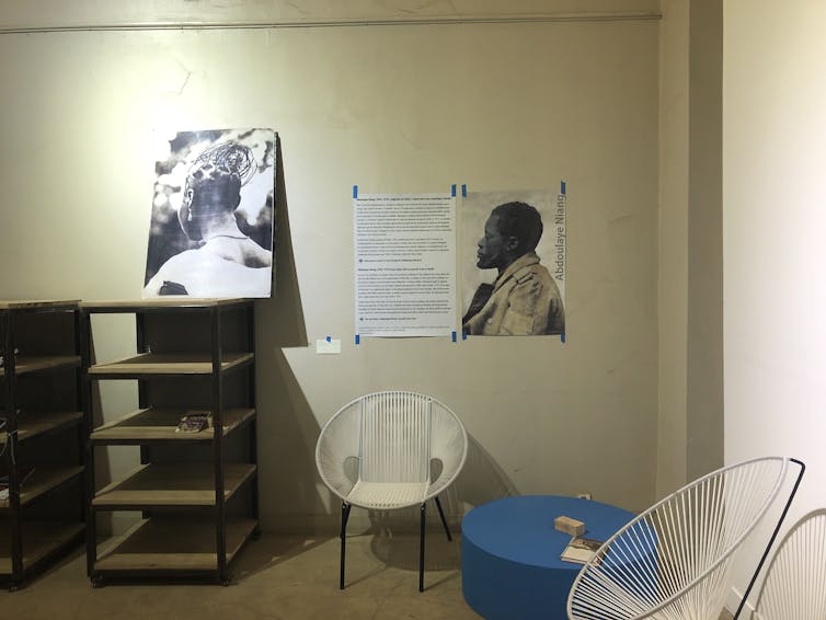 A room with chairs and shelves and a table. On the wall, a poster of a portrait of an African man and on the top of the shelves a photo of an African woman from behind, hair braided elaborately.