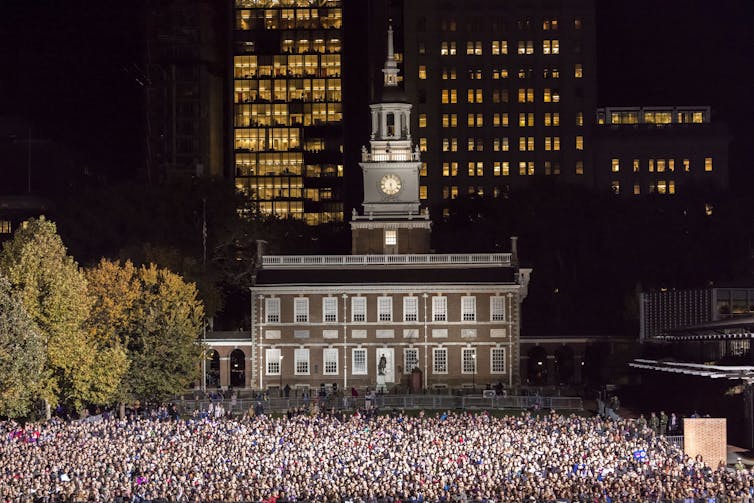 A crowd of thousands gathered in the nighttime in front of a historic building with a bell tower.