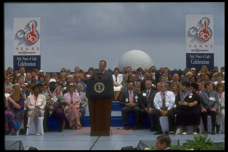 A man in a suit speaking at a lectern with a crowd behind him.