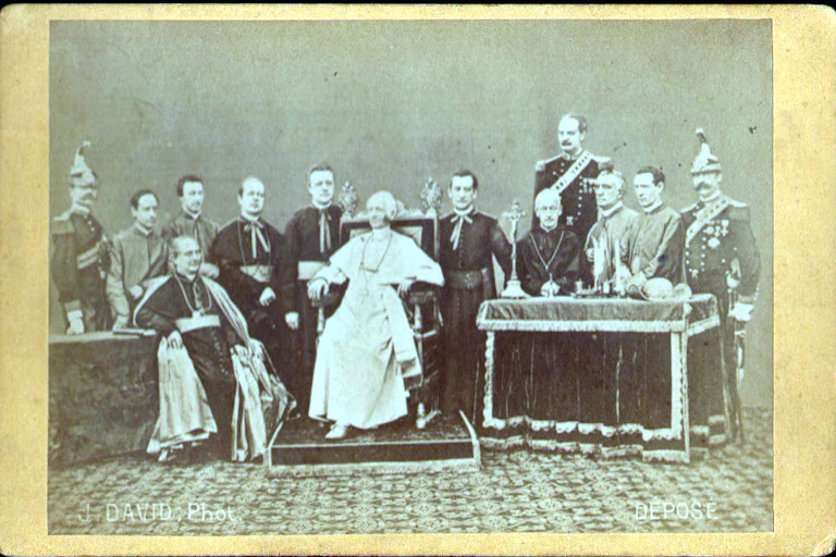 A faded, black and white photograph of a handful of men formally posed around a man dressed in white who sits on a throne.