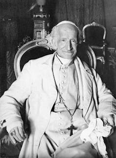 A black and white photo of a smiling man in white robes, wearing a heavy cross necklace.