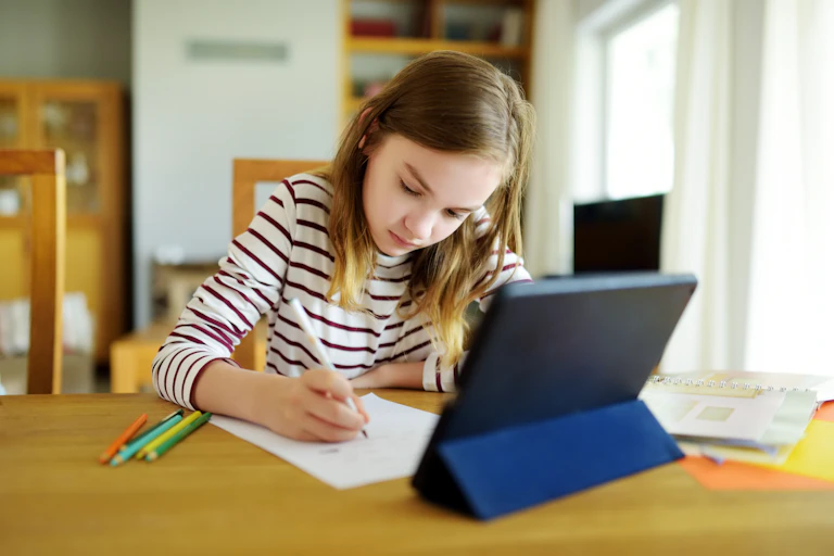 A young girl writes on a sheet of paper at a desk, in front of a tablet.