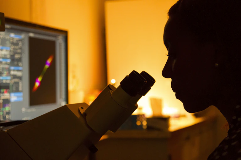 A woman looking into a microscope next to a computer.
