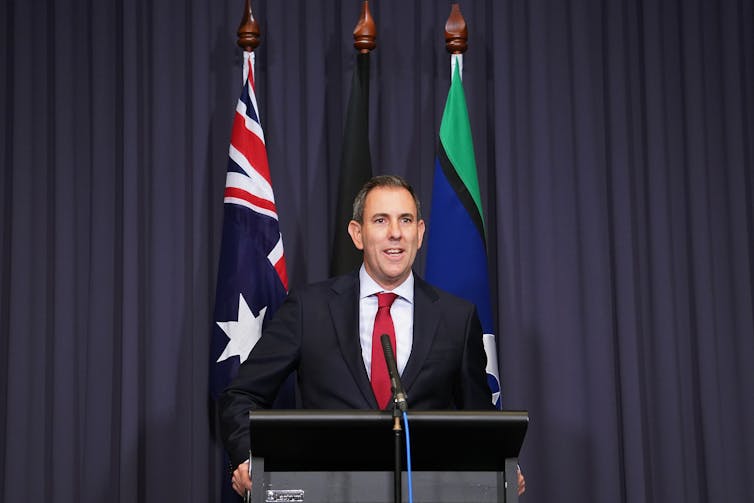 A man wearing a black suit and a red tie speaking at a microphone.