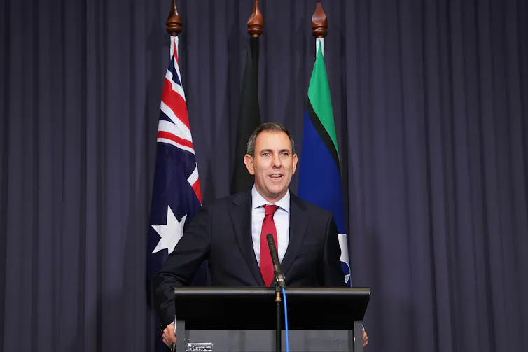 A man wearing a black suit and a red tie speaking at a microphone.