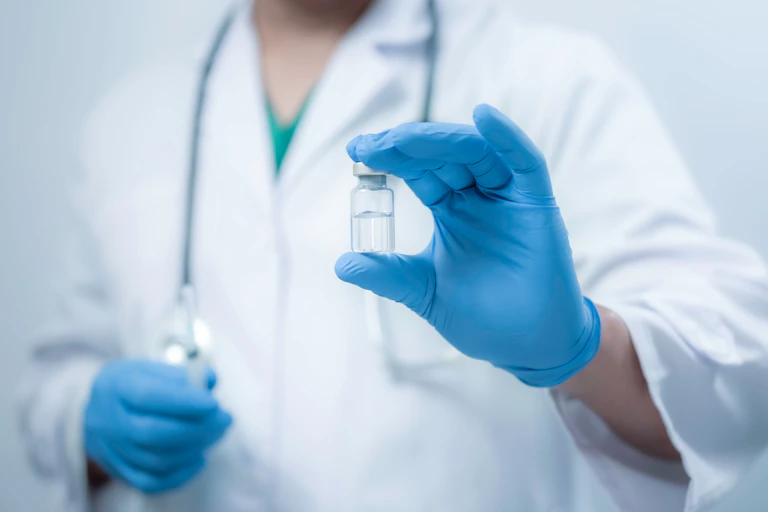 A person in a white lab coat holding up an unmarked phial containing a clear liquid.