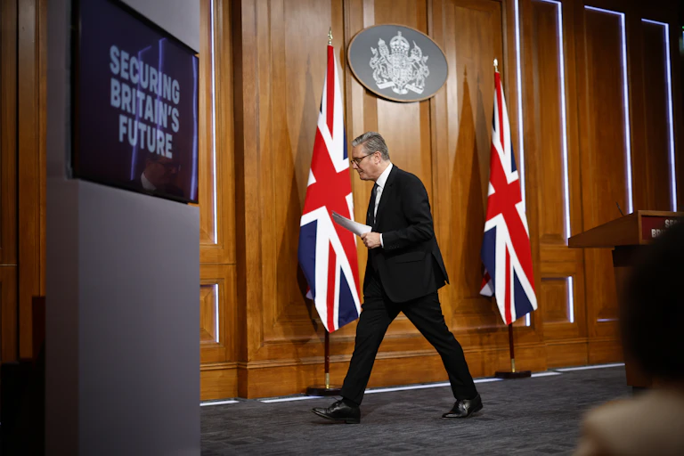 Keir Starmer walking away from a podium, past two UK flags and toward a screen reading 'securing britain's future'