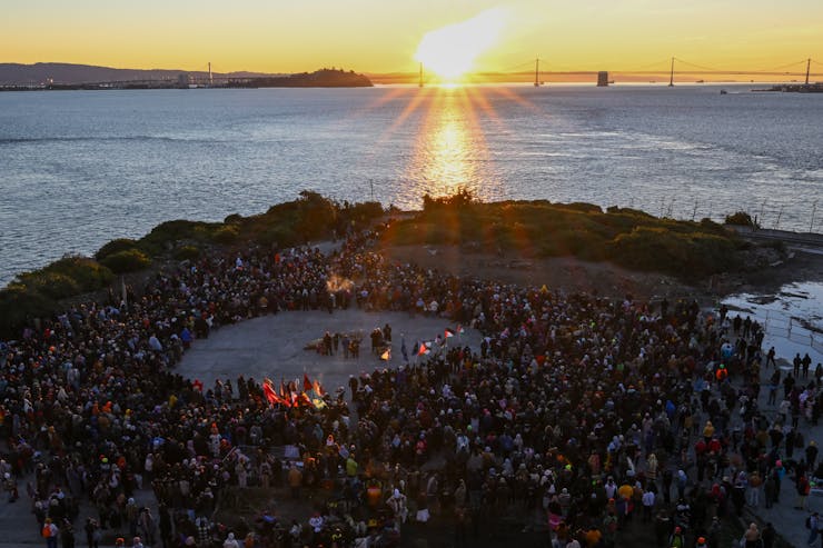 A group of people stand around an open circle on an island, as the Sun rises behind a bridge across the water.