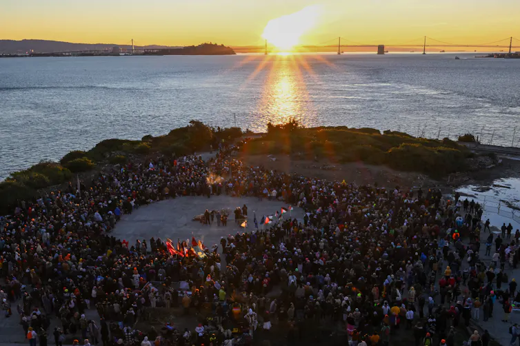 A group of people stand around an open circle on an island, as the Sun rises behind a bridge across the water.