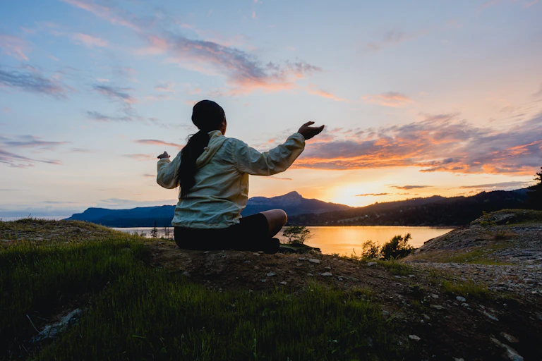 A person sits on the ground, raising her arms to greet the rising Sun, emerging behind distant hills and water.