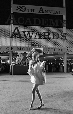 Woman holds paper over her head to protect her hair as she walks across a carpet wearing high heels and a short skirt.