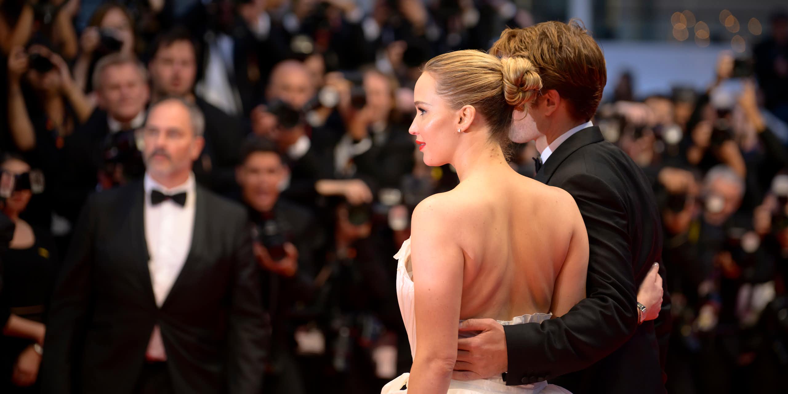 Young man wearing tuxedo poses with young woman wearing white gown before a large group of photographers.