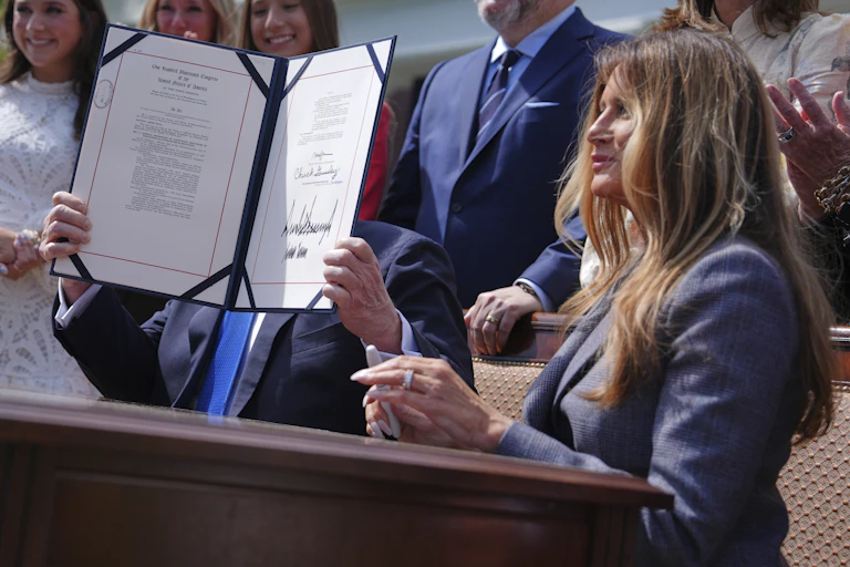 a seated man in a business suit holds up a document, obscuring his face, while seated next to a woman outdoors with several people standing behind them