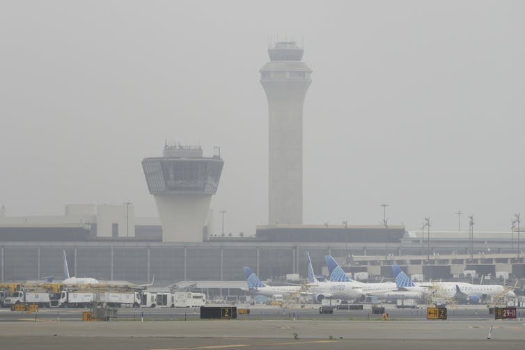 Two towers with big glass windows stand over airplanes sitting on the tarmac in this foggy scene